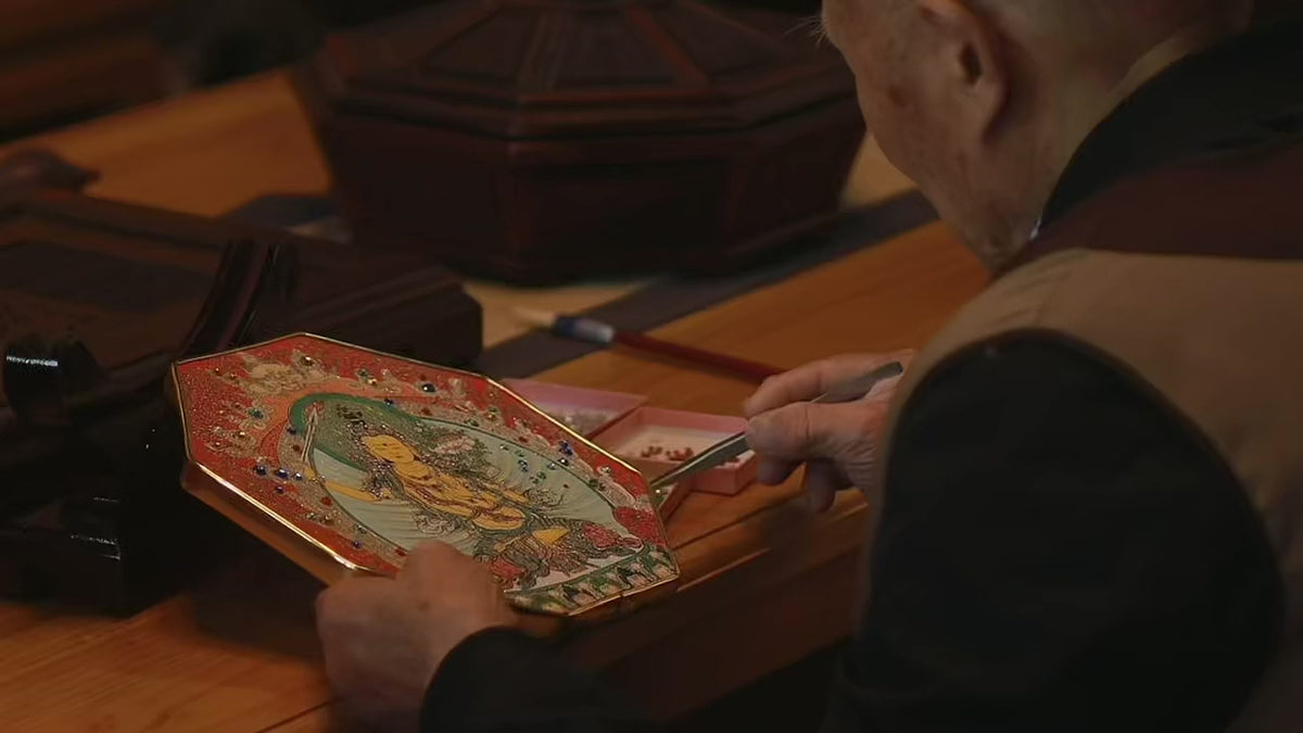 At the temple, an elderly lay Buddhist sets crystals into a thangka by hand with tweezers